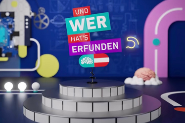 Und wer hat's erfunden? Eine Bühne mit gefliesten Stufen steht mittig vor einem farbenfrohen, abstrakten Hintergrund mit Wissenschaftsmotiven. Auf Schildern über der Bühne steht: „UND WER HATS ERFUNDEN?“ Rechts sitzt ein Gehirnmodell.