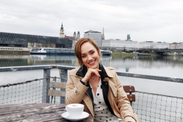 Reisezeit Eine lächelnde Frau in einem beigen Trenchcoat sitzt an einem bewölkten Tag mit einem Cappuccino an einem Cafétisch im Freien, im Hintergrund sind ein Fluss und Stadtgebäude zu sehen.